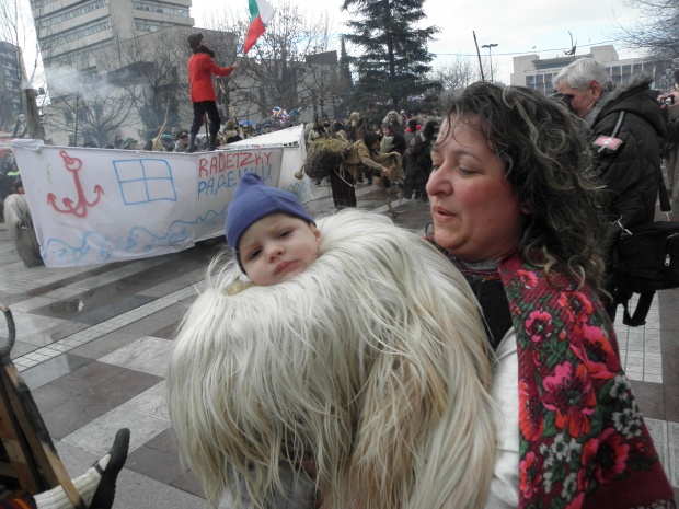 Вероятно най-малкият участник в традиционното шествие на кукери и сурвакари в Благоевград се е сгушил в костюма си в ръцете на мама.